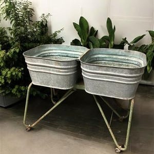 May include: A galvanized metal planter with two rectangular tubs. The tubs are supported by a light green metal frame with wheels. Green plants are visible in the background.