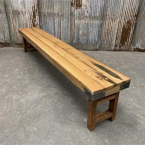 May include: A long, rectangular wooden bench with a natural finish. The bench is constructed from several planks of wood and features metal corner brackets. The legs are made of wood and are a darker shade than the bench top. The bench measures approximately 72 inches long.