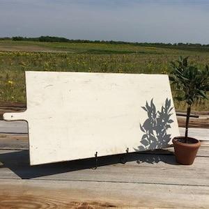 Puede incluir: Una tabla de cortar rectangular grande de color blanco con un asa en el lado izquierdo. La tabla está de pie sobre una superficie de madera con una pequeña planta en maceta en primer plano.