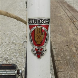 Op de afbeelding: Close-up van een vintage fietsframe met een witte afwerking. Het frame heeft een "Rudge"-embleem met een rood en zwart ontwerp, inclusief een handsymbool en de tekst "Nottingham England".