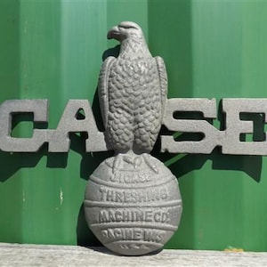 May include: A gray metal sculpture featuring an eagle perched atop a globe, with the word "CASE" in large block letters. The globe has text that reads "CASE THRESHING MACHINE CO. RACINE WIS."