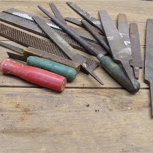 May include: A collection of metal files and rasps with wooden handles, some with green and red handles, on a wooden surface.