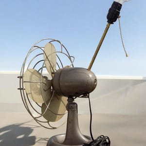 Puede incluir: Un ventilador vintage de metal con una bombilla en la parte superior. El ventilador tiene una base de metal gris y un cable negro.