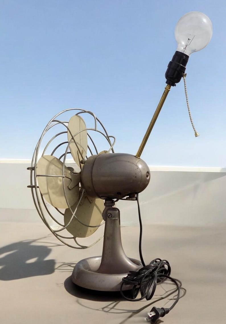 Puede incluir: Un ventilador vintage de metal con una bombilla en la parte superior. El ventilador tiene una base de metal gris y un cable negro.