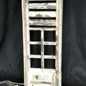 May include: A tall, rectangular wooden window shutter with a distressed white and grey paint finish. The upper section features horizontal slats, while the lower section is divided into six small square panes. A sprig of white artificial flowers.