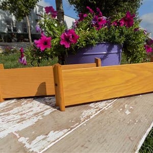 May include: Two wooden rectangular planters with a golden-brown finish are displayed outdoors. The planters are empty and ready for planting. A purple bucket filled with vibrant pink flowers sits in the background, adding a pop of color to the scene.