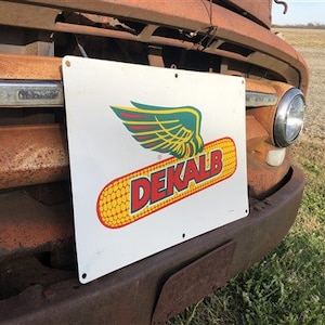Op de afbeelding: Een vintage wit metalen bord met een rood, geel en groen logo voor DeKalb. Het logo toont een gestileerde vleugel en een kolf maïs. Het bord is bevestigd aan de voorkant van een roestige, bruine vrachtwagen.