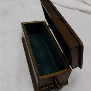 May include: A wooden box with a hinged lid and a small knob on the front. The box is lined with green velvet and has a decorative border around the edges.