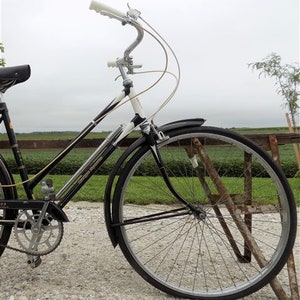 Op de afbeelding: Een vintage zwart-witte fiets met een gebogen stuur en een zwart zadel. De fiets heeft een zilveren ketting en zwarte banden. De fiets staat op een grindoppervlak.