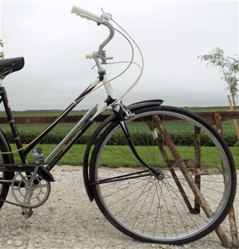 Op de afbeelding: Een vintage zwart-witte fiets met een gebogen stuur en een zwart zadel. De fiets heeft een zilveren ketting en zwarte banden. De fiets staat op een grindoppervlak.