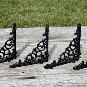 Peut inclure: Quatre supports d'étagère en métal noir avec un motif de volutes décoratif. Les supports sont disposés en rangée sur une surface en bois.