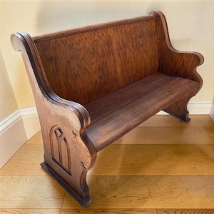 May include: A medium-brown wooden church pew featuring a high backrest with a visible wood grain pattern, curved armrests, and side panels decorated with a carved arched window design. The bench has a solid seat and sturdy base construction.