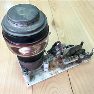 Op de afbeelding: Een antiek elektronisch onderdeel met een cilindrische, meerlaagse structuur, een metalen bovenkant en een glazen gedeelte dat koperdraad laat zien. Het apparaat is gemonteerd op een rechthoekige basis met diverse kleine mechanische onderdelen en bedrading.