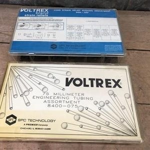 Puede incluir: Dos contenedores de plástico transparente con información del producto. El contenedor superior es azul y blanco, etiquetado como "VOLTREX brand strain reliefs". El contenedor inferior es beige, con "VOLTREX" y "75 MILLIMETER ENGINEERING TUBING ASSORTMENT".