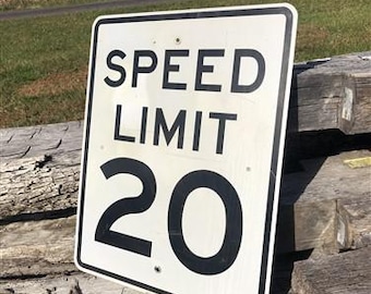 Peut inclure: Panneau de limitation de vitesse rectangulaire blanc avec texte noir et bordure noire. Le panneau indique « SPEED LIMIT 20 ». Le panneau est monté sur une surface en bois vieilli, avec un paysage rural en arrière-plan.
