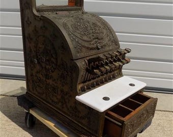 Könnte beinhalten: Antike Registrierkasse mit aufwendigen floralen Details und einer weißen Marmorablage. Die Kasse ist aus dunklem Metall gefertigt und steht auf einer rollbaren Plattform. Das Wort "SALE" ist oben sichtbar.