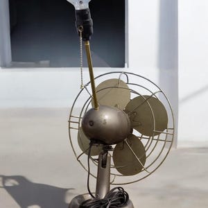 Puede incluir: Un ventilador vintage de metal con una bombilla en la parte superior. El ventilador tiene tres aspas y una jaula de metal. La bombilla es transparente y tiene un filamento de metal.