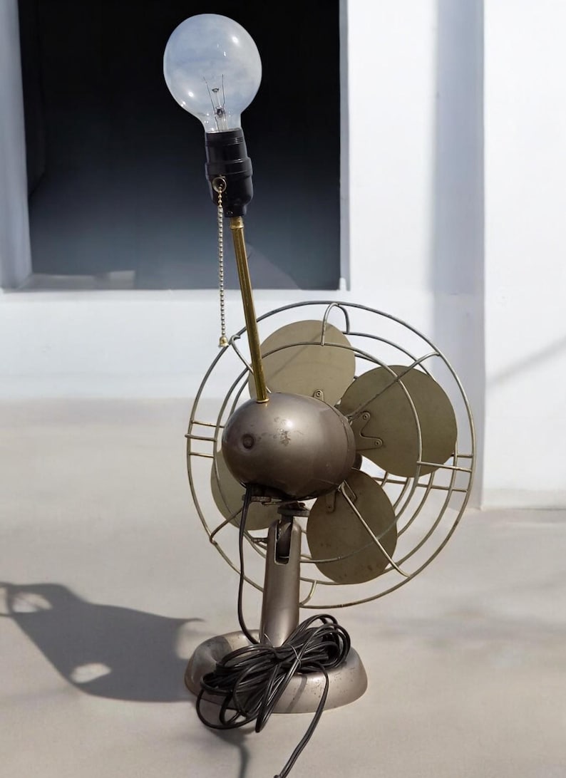 Puede incluir: Un ventilador vintage de metal con una bombilla en la parte superior. El ventilador tiene tres aspas y una jaula de metal. La bombilla es transparente y tiene un filamento de metal.