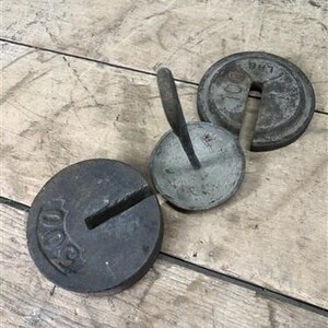 Puede incluir: Tres pesas antiguas de hierro fundido sobre una superficie de madera. Dos son redondas, marcadas "100 LBS", y una es una pesa circular más pequeña con un asa. Las pesas son de color gris oscuro y muestran signos de desgaste.