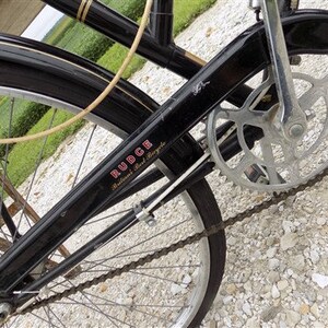 Op de afbeelding: Close-up van een vintage zwarte fiets met zilveren ketting en tandwielen. De fiets heeft een zwart frame met het woord "RUDGE" in rode letters. De banden zijn zwart en de spaken zijn zilver.