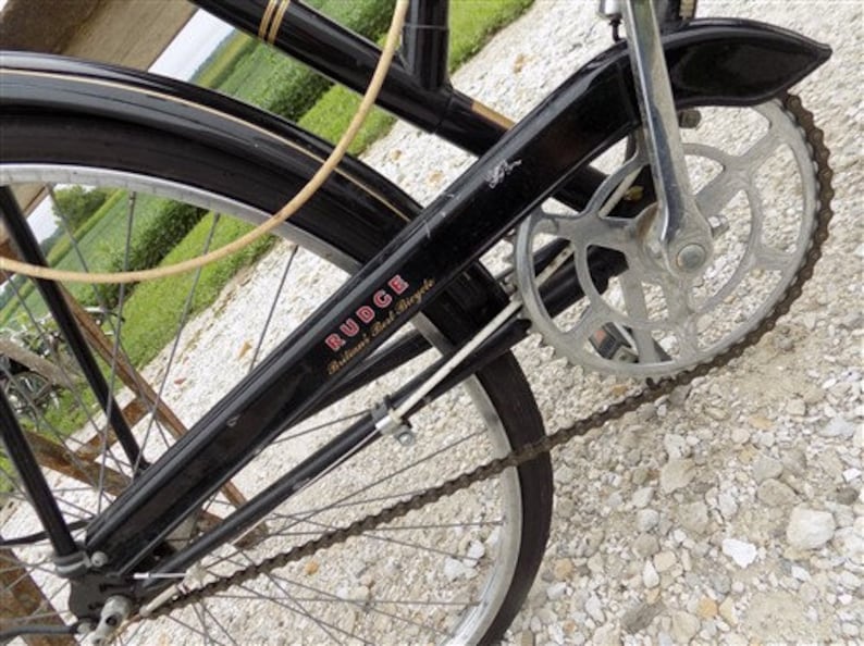 Op de afbeelding: Close-up van een vintage zwarte fiets met zilveren ketting en tandwielen. De fiets heeft een zwart frame met het woord "RUDGE" in rode letters. De banden zijn zwart en de spaken zijn zilver.