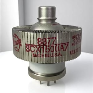 Puede incluir: Un tubo de vacío plateado con un diseño cilíndrico. El tubo tiene una estructura circular con aletas con texto rojo que dice "8877 3CX1500A7 MADE IN U.S.A." La base es blanca con varios pines.