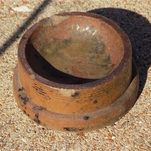 May include: A small, round, terracotta-colored bowl with a black rim and a worn, textured surface. The bowl has a slightly raised base.