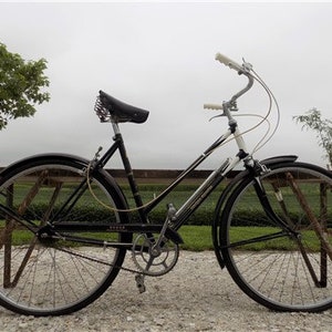 Op de afbeelding: Een vintage zwarte fiets met zilveren accenten, een gebogen stuur en een klassiek leren zadel. De fiets heeft zwarte spatborden en banden en staat op een grindoppervlak. Een klassieke fiets.