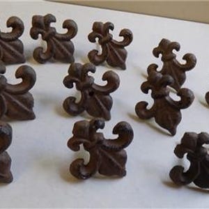 May include: A collection of eleven brown fleur-de-lis cabinet knobs. The knobs are made of metal and have a detailed, ornate design. They are arranged on a white surface.