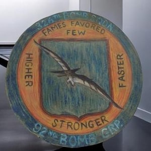 Puede incluir: Un letrero redondo pintado de naranja y azul con un pterodáctilo volando en el centro. El letrero dice "326th Bomb Squadron, Fames Favored Few, Higher, Faster, Stronger, 92nd Bomb Grp."