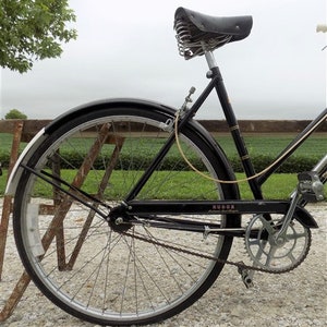 Op de afbeelding: Zwarte vintage fiets met een leren zadel en een zwart frame. De fiets heeft een zilveren ketting en een zilveren tandwiel. De banden zijn zwart met zilveren velgen. De fiets staat op een grindoppervlak.