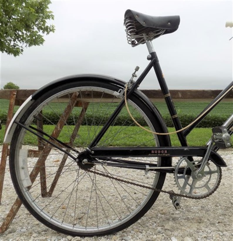 Op de afbeelding: Zwarte vintage fiets met een leren zadel en een zwart frame. De fiets heeft een zilveren ketting en een zilveren tandwiel. De banden zijn zwart met zilveren velgen. De fiets staat op een grindoppervlak.