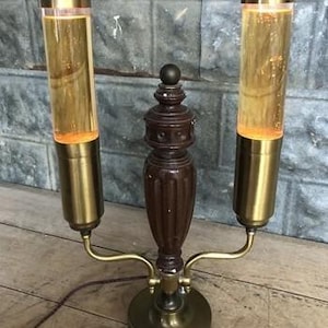 Könnte beinhalten: Eine Vintage-Tischlampe mit zwei zylindrischen Glasröhren, die mit bernsteinfarbener Flüssigkeit gefüllt sind. Die Lampe hat eine dunkelbraune Mittelsäule aus Holz und messingfarbene Metallakzente. Der Sockel ist rund und die Lampe ist eingesteckt.