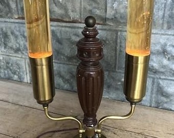 Puede incluir: Una lámpara de mesa vintage con dos tubos de vidrio cilíndricos llenos de líquido ámbar. La lámpara tiene una columna central de madera marrón oscuro y detalles metálicos de color latón. La base es redonda y la lámpara está enchufada.
