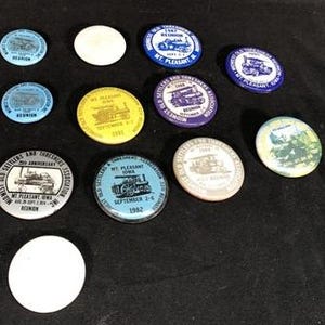Può includere: Una collezione di 11 spille rotonde con vari disegni. Le spille presentano immagini di motori a vapore, treni e altri veicoli storici. Le spille sono di vari colori, tra cui blu, rosso, giallo e verde. Il testo sulle spille include "Old Settlers and Thresher Association", "Mt. Pleasant, Iowa" e "1982".