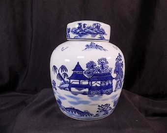Puede incluir: Un jarrón de cerámica blanca con un diseño azul y blanco de un paisaje chino. El jarrón tiene una tapa y está decorado con una pagoda, árboles y agua.