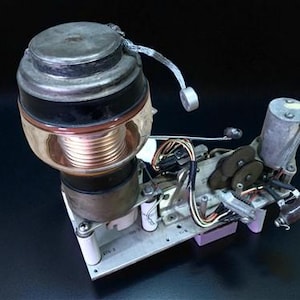 Puede incluir: Un dispositivo mecánico complejo con una bobina de cobre cilíndrica, una carcasa de vidrio y varios componentes metálicos. El dispositivo tiene una parte superior e inferior plateadas, con detalles en negro y marrón. Se ven cables y engranajes.