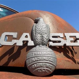 May include: Close-up of a vintage rusty truck hood with the word "CASE" in large silver letters. A detailed silver eagle emblem sits atop a globe-shaped base. The truck's hood is a weathered reddish-brown color, with a clear blue sky in the background.