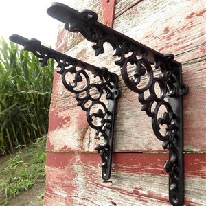 Peut inclure: Deux supports d'étagère en fonte noire avec un motif de volutes décoratif. Les supports sont fixés sur une surface en bois vieilli.