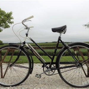 Op de afbeelding: Een vintage zwarte fiets met witte handvatten en een zwart zadel. De fiets heeft zilveren velgen en spatborden. De fiets staat geparkeerd op een grindoppervlak met een veld en bomen op de achtergrond.
