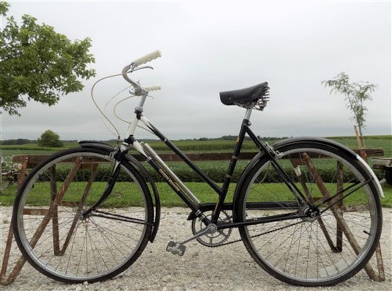 Op de afbeelding: Een vintage zwarte fiets met witte handvatten en een zwart zadel. De fiets heeft zilveren velgen en spatborden. De fiets staat geparkeerd op een grindoppervlak met een veld en bomen op de achtergrond.