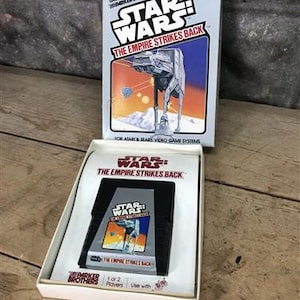 May include: A vintage Star Wars: The Empire Strikes Back video game cartridge for Atari and Sears systems. The black cartridge sits in a white cardboard tray, accompanied by its original rectangular box featuring an AT-AT walker illustration.