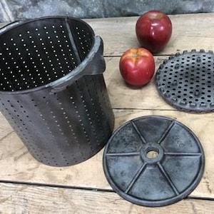 May include: An antique apple press with a perforated metal bucket, two round metal plates, and two red apples. The bucket has handles and is cylindrical. The plates are circular with holes. The apples are red.