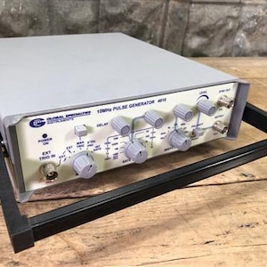 Puede incluir: Un generador de impulsos de 10 MHz gris con múltiples perillas y diales. El panel frontal tiene el texto "GLOBAL SPECIALTIES INSTRUMENTS" y "10MHz PULSE GENERATOR 4010". Tiene un asa negra y está sobre una superficie de madera.