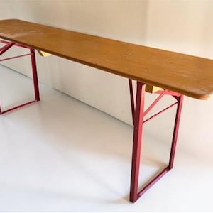 May include: A long, rectangular wooden table with a light brown surface. The table has red metal legs and support beams. The table is approximately 96 inches long.