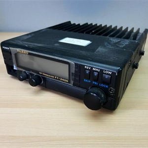 May include: A black Yaesu FT-2400 FM transceiver with a digital display and various buttons. The device has a series of cooling fins on top and is set on a light-colored wooden surface. The text "Yaesu" is visible on the front.
