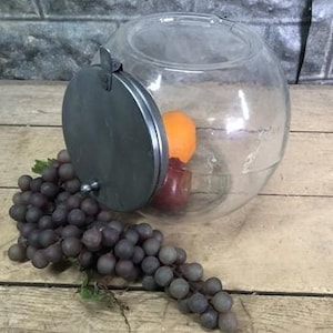 Pode incluir: Um frasco de vidro transparente com uma tampa de metal e uma trava com dobradiça. O frasco está cheio de frutas, incluindo uma laranja e uma maçã. Um cacho de uvas está em primeiro plano.