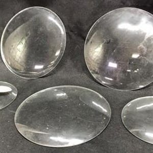Può includere: Cinque lenti convesse trasparenti di varie dimensioni. Due sono grandi e circolari, mentre le altre sono più piccole e ovali. Le lenti sono trasparenti e riflettono la luce, suggerendo che siano fatte di vetro o materiale simile.