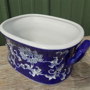 Peut inclure: Un pot de fleurs en céramique bleu et blanc avec une poignée. Le pot a un intérieur blanc et un extérieur bleu avec un motif floral. Le pot est de forme ovale et repose sur une surface en bois.