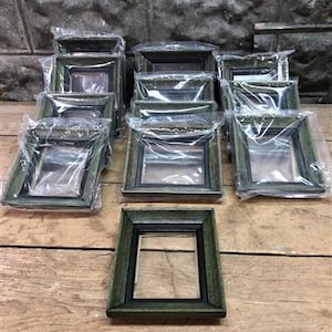 以下が含まれることがあります： 黒のマットが付いた緑色の写真フレームが10個セットです。フレームは木でできており、使い古したような仕上げになっています。すべて同じサイズで、木製の表面に一列に並べてあります。